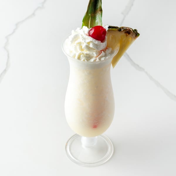 Creamy white cocktail in a tall glass, topped with whipped cream, a cherry, and pineapple.