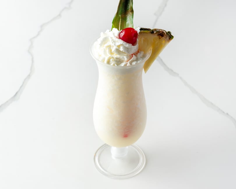 Creamy white cocktail in a tall glass, topped with whipped cream, a cherry, and pineapple.