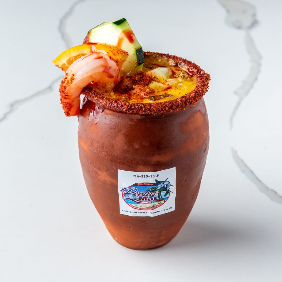 A Perlas Del Mar michelada in a clay cup, garnished with shrimp, cucumber, and orange.