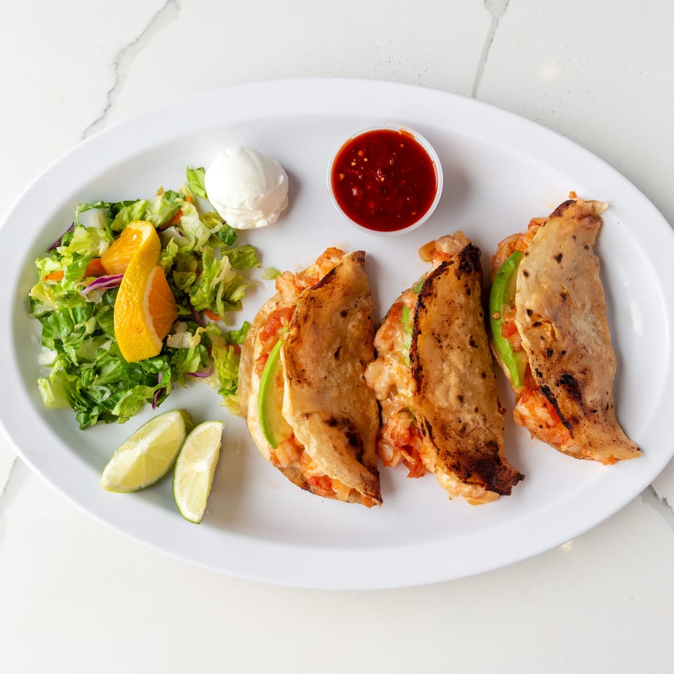 Three shrimp tacos with avocado, a side salad, lime wedges, sour cream, and salsa.