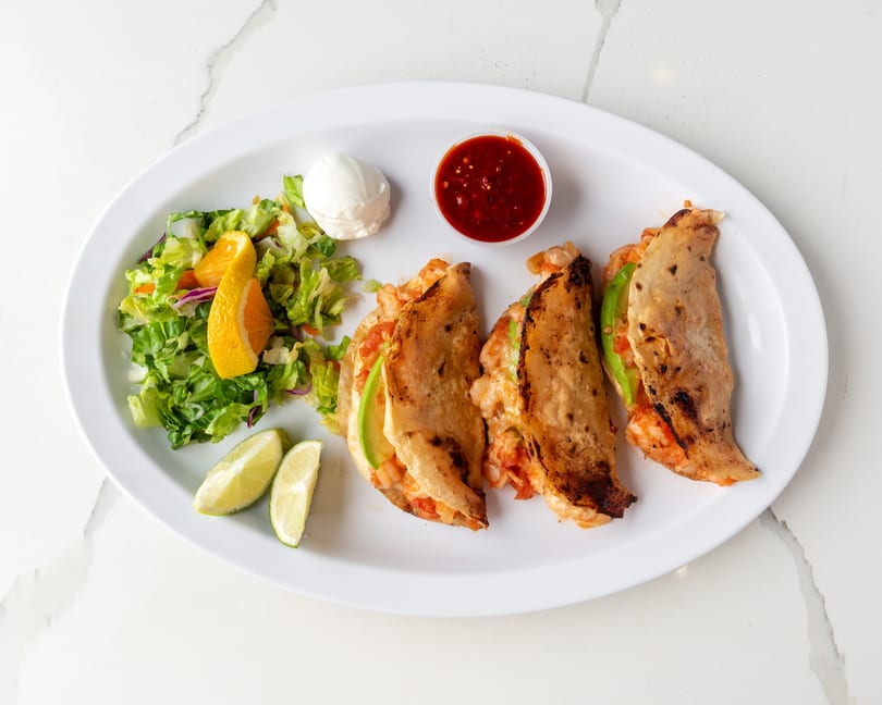 Three shrimp tacos with avocado, a side salad, lime wedges, sour cream, and salsa.