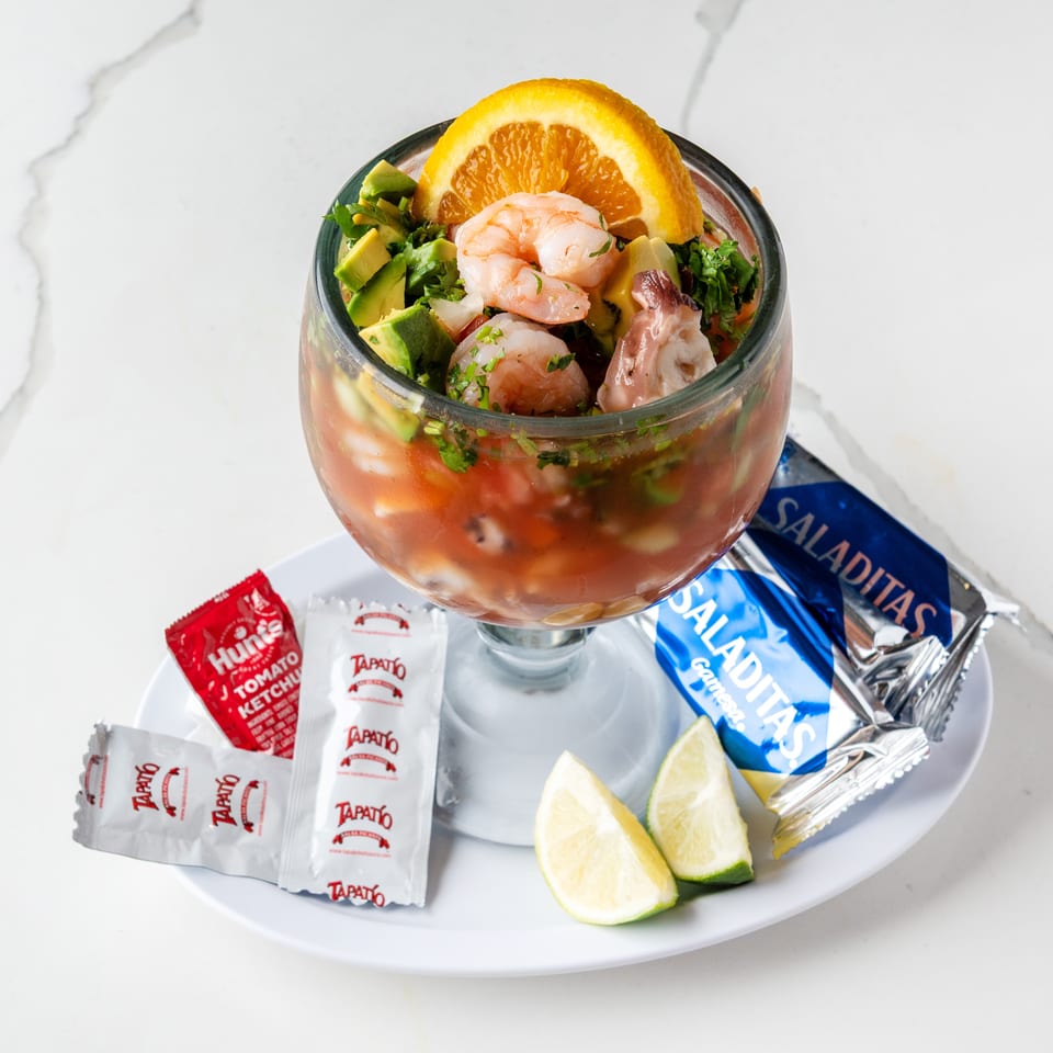 Shrimp and avocado ceviche in a glass, with lime wedges, hot sauce, ketchup, and crackers.
