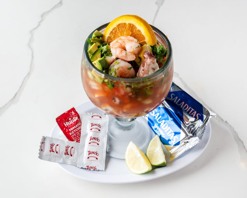 Shrimp and avocado ceviche in a glass, with lime wedges, hot sauce, ketchup, and crackers.