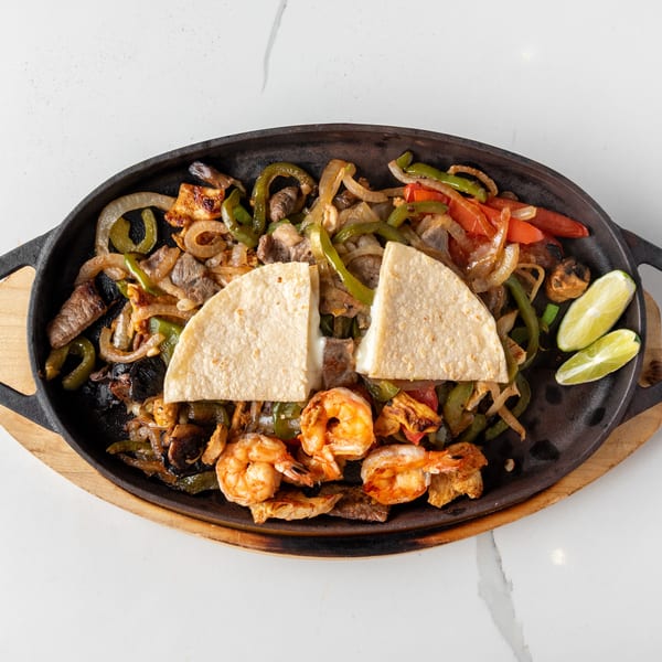 Sizzling fajitas with shrimp, steak, chicken, peppers, onions, and tortillas with lime wedges.