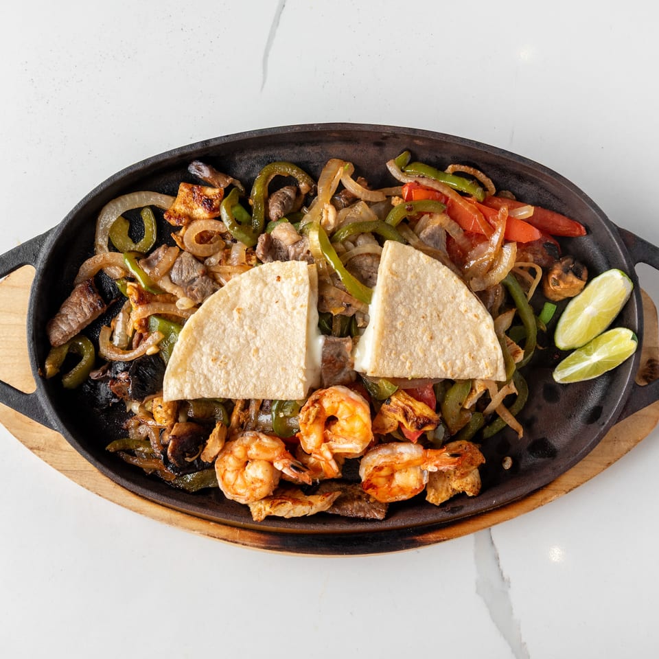Sizzling fajitas with shrimp, steak, chicken, peppers, onions, and tortillas with lime wedges.