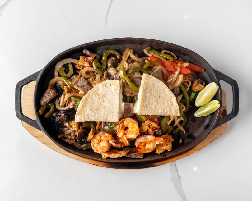 Sizzling fajitas with shrimp, steak, chicken, peppers, onions, and tortillas with lime wedges.