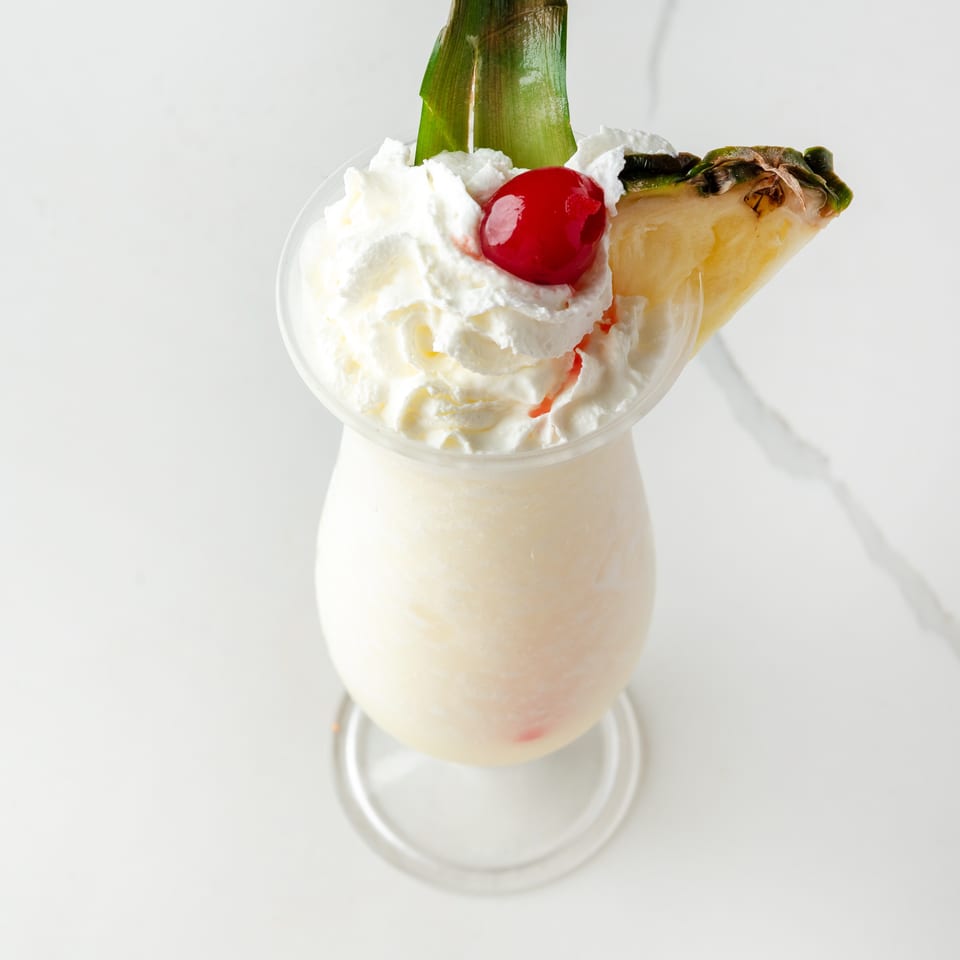 Creamy white cocktail with whipped cream, a cherry, pineapple wedge, and green leaf garnish.