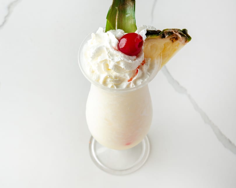 Creamy white cocktail with whipped cream, a cherry, pineapple wedge, and green leaf garnish.