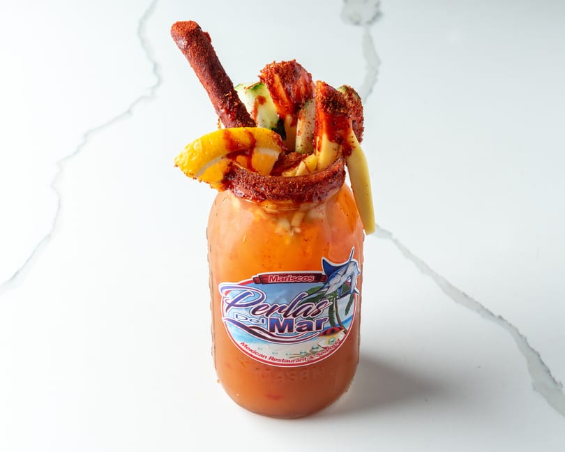 A Perlas Del Mar Michelada, garnished with cucumber, mango, lemon, and a tamarind candy straw.