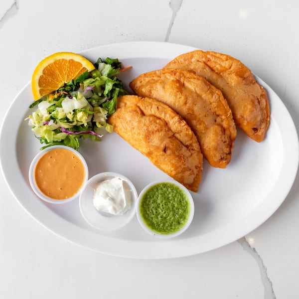 Three golden-brown empanadas, a side salad with orange slice, and three dipping sauces on a white plate.