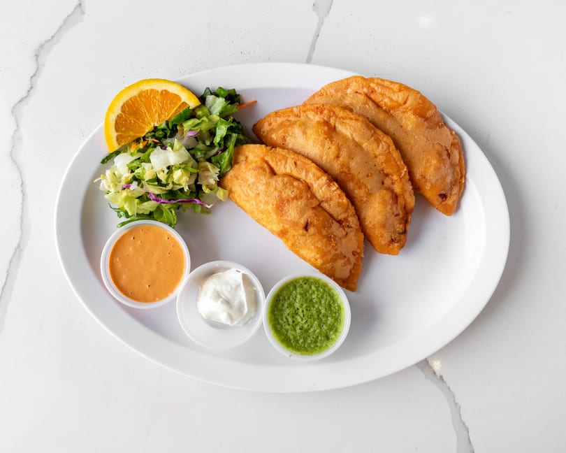 Three golden-brown empanadas, a side salad with orange slice, and three dipping sauces on a white plate.
