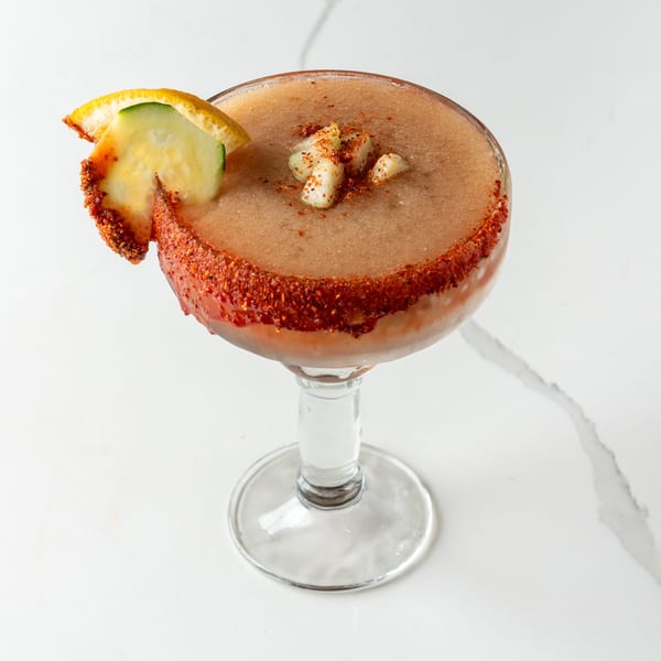 A frozen cocktail with a chili-lime rim, garnished with cucumber and orange slices.
