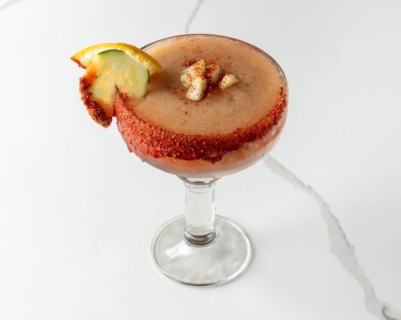 A frozen cocktail with a chili-lime rim, garnished with cucumber and orange slices.