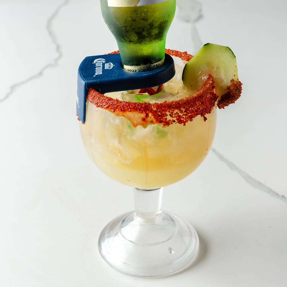 Michelada cocktail in a goblet with a Corona beer bottle, chili-lime rim, and cucumber slice.