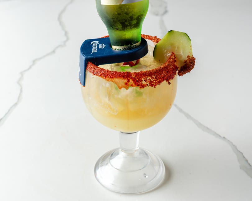 Michelada cocktail in a goblet with a Corona beer bottle, chili-lime rim, and cucumber slice.