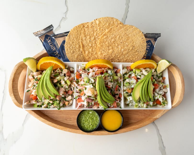 Three types of ceviche with avocado, orange, and lime, served with tostadas and sauces.