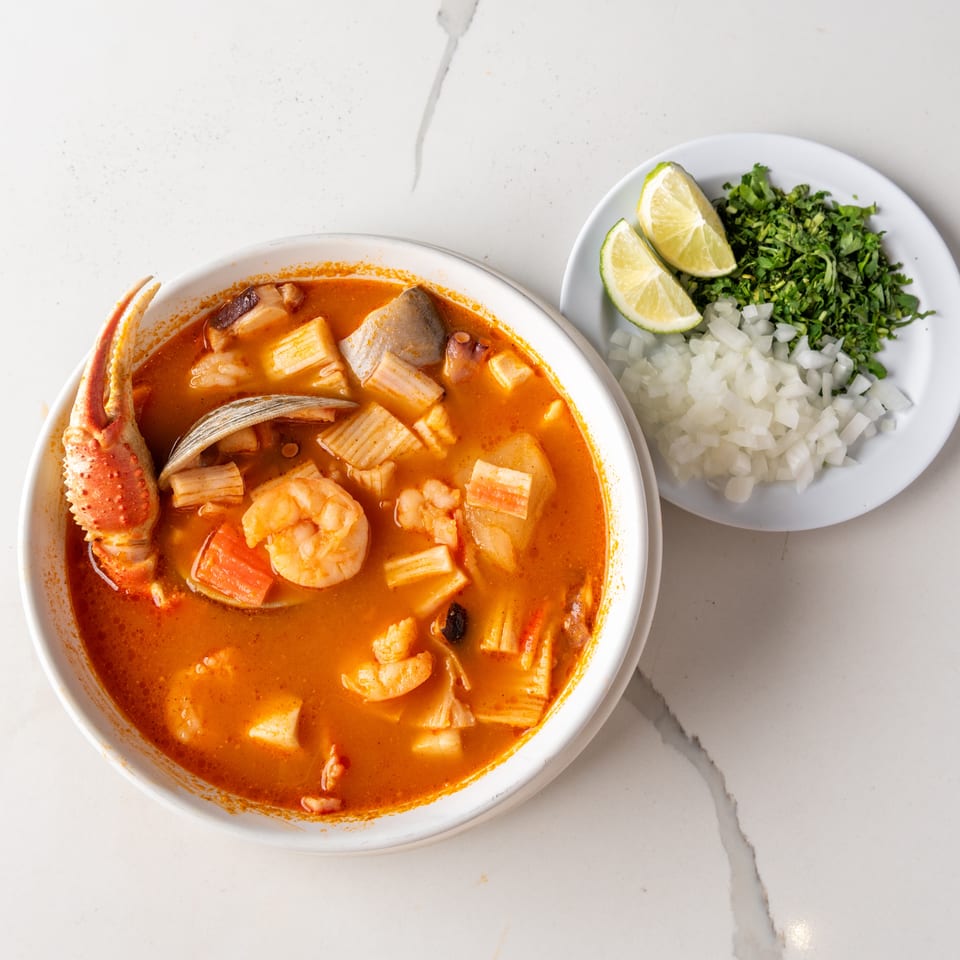 A vibrant seafood soup with crab, shrimp, and vegetables, served with a side of chopped onions, cilantro, and lime.