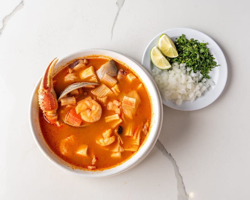 A vibrant seafood soup with crab, shrimp, and vegetables, served with a side of chopped onions, cilantro, and lime.