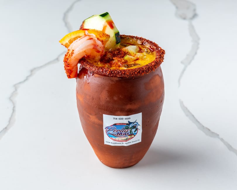 A Perlas Del Mar michelada in a clay cup, garnished with shrimp, cucumber, and orange.