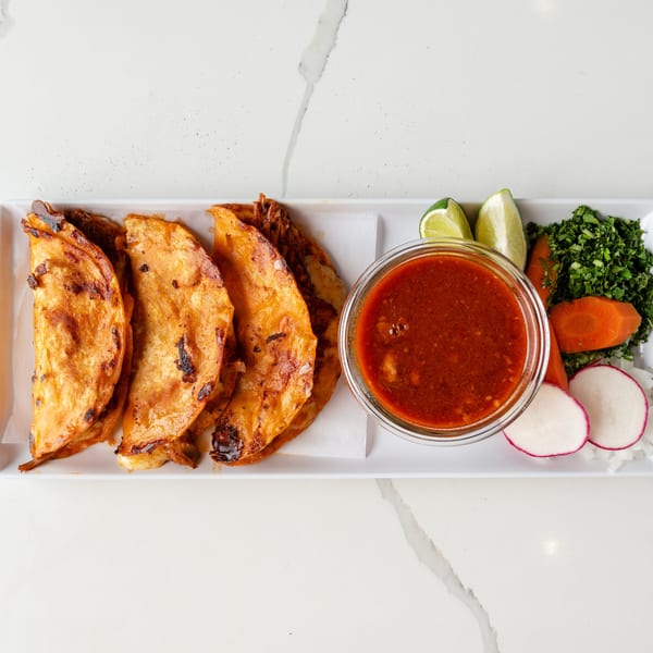 Four crispy birria tacos with a side of consommé, lime wedges, cilantro, carrots, radishes, and onions.