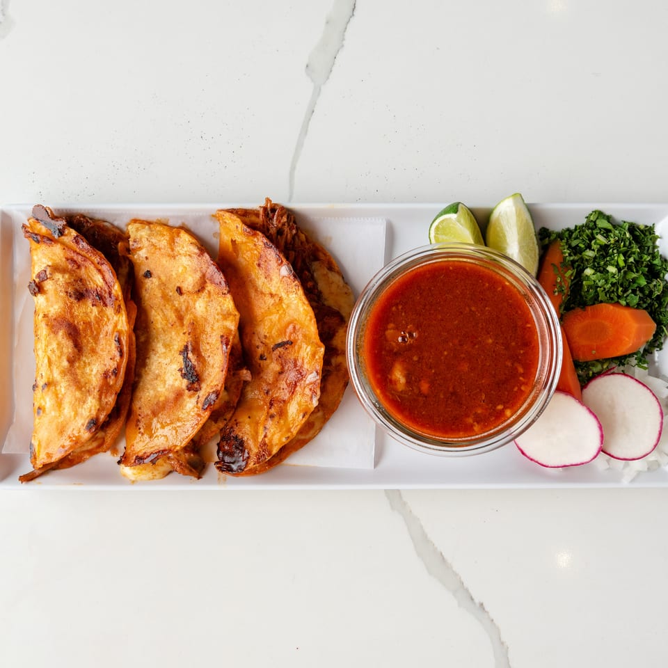 Four crispy birria tacos with a side of consommé, lime wedges, cilantro, carrots, radishes, and onions.