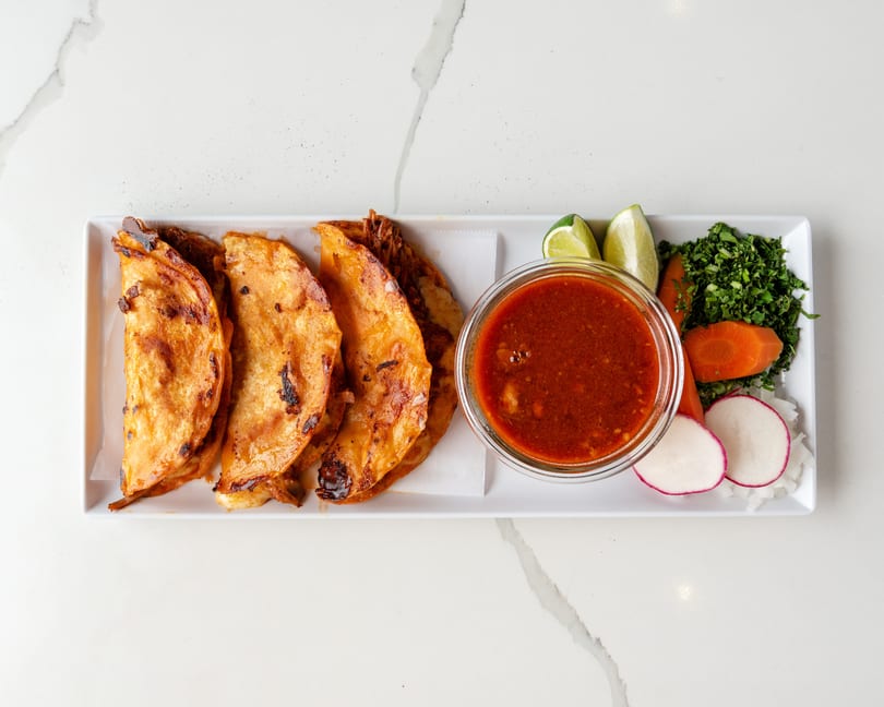 Four crispy birria tacos with a side of consommé, lime wedges, cilantro, carrots, radishes, and onions.