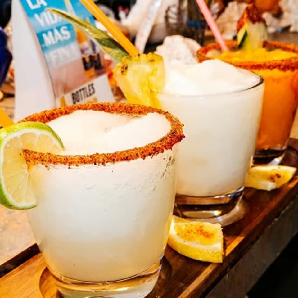 Three frozen margaritas, two white and one orange, garnished with lime, pineapple, and cucumber.