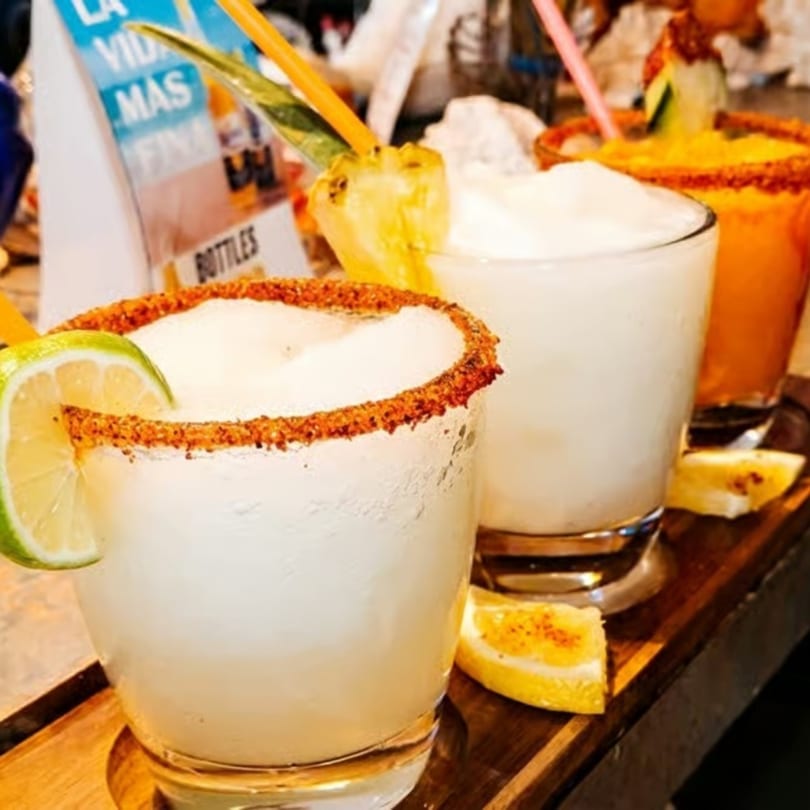 Three frozen margaritas, two white and one orange, garnished with lime, pineapple, and cucumber.